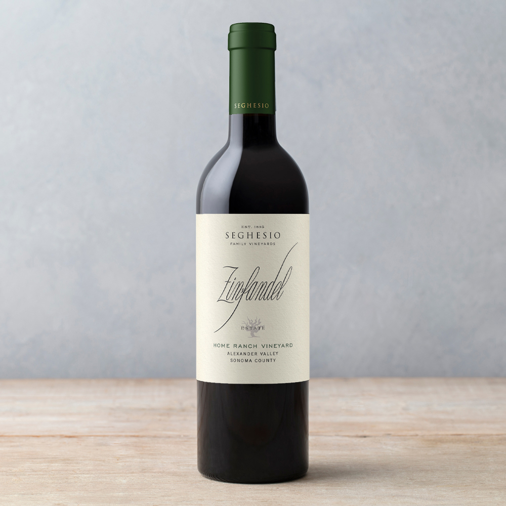 A bottle of Seghesio Family Vineyards Home Ranch Zinfandel wine with a green foil top sits on a light wooden surface against a plain, light gray background.