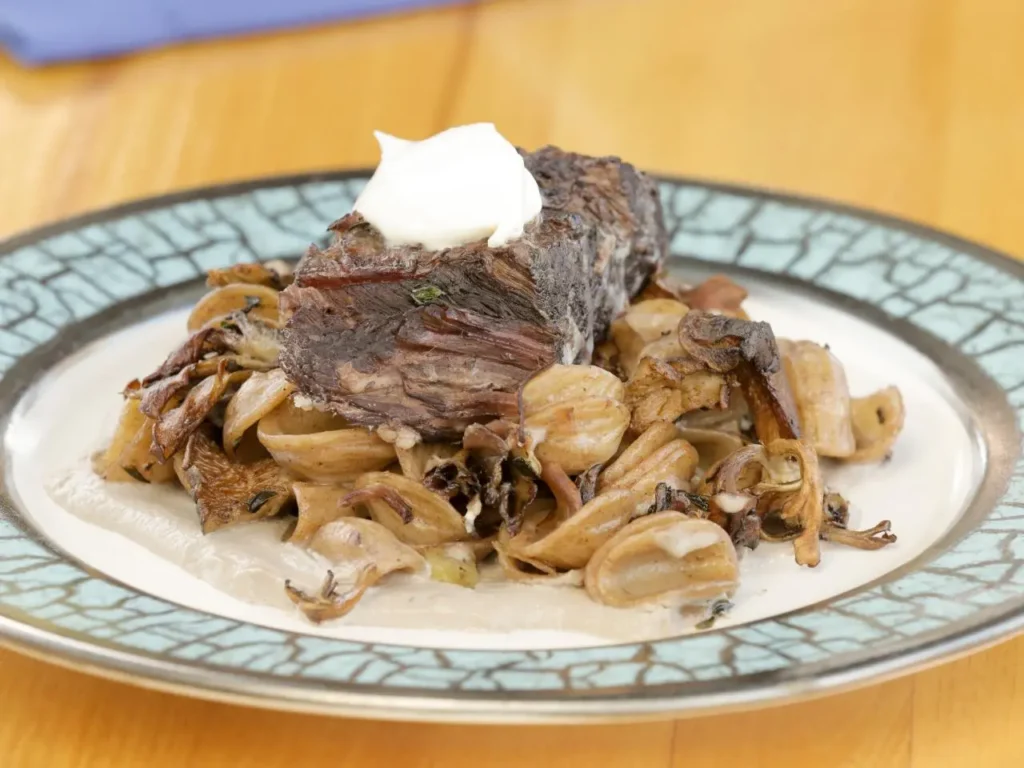 A plate with braised beef topped with a dollop of sour cream, served on a bed of sautéed mushrooms and onions, all on a creamy sauce. The dish is presented on a patterned plate.