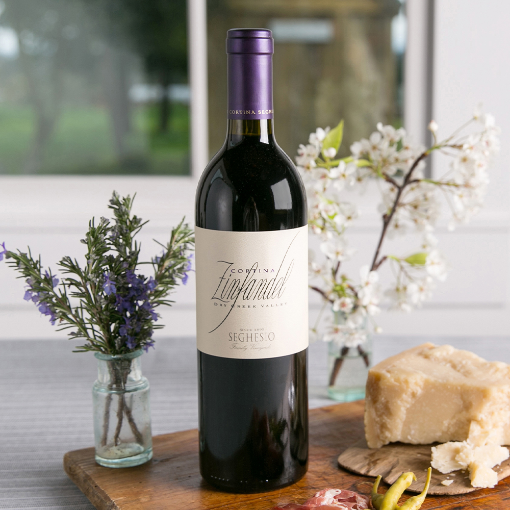 Cortina Zinfandel wine bottle on wood cutting board with flowers and a block of cheese beside