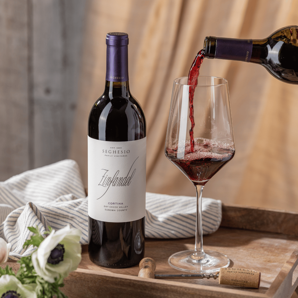 A bottle of Seghesio Zinfandel wine stands next to a glass being filled with red wine on a wooden tray, with a striped cloth, a cork, and white flowers nearby.