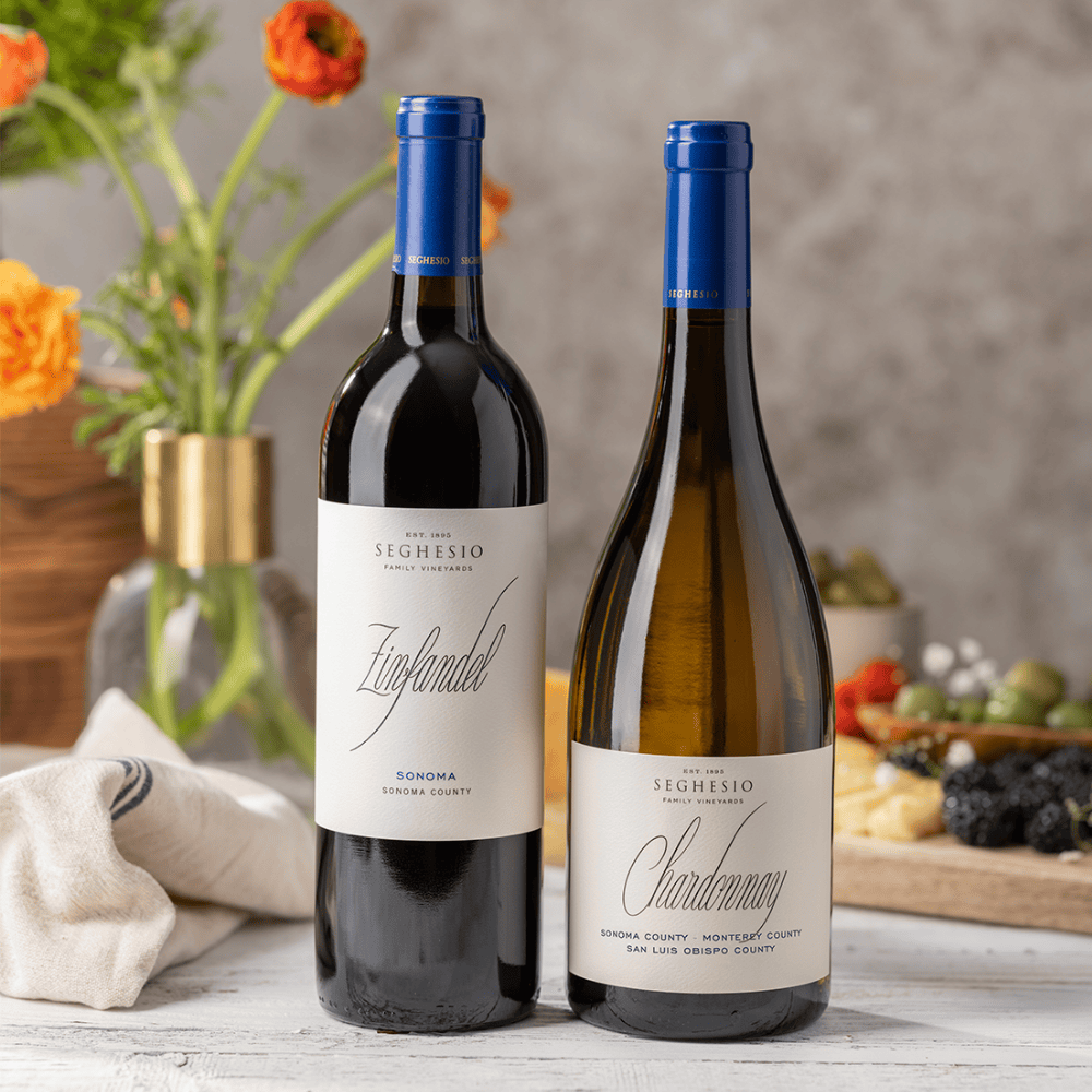 Two bottles of Seghesio wine, a Zinfandel and a Chardonnay, stand side by side on a table with a white cloth, flowers in a vase, and a blurred cheese and fruit platter in the background.