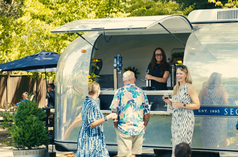 Three people stand and chat outside a shiny silver food trailer while a smiling server stands inside. It’s a sunny day with trees and patio umbrellas in the background. The scene looks cheerful and relaxed.