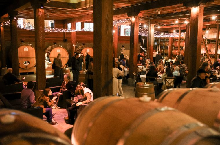 People gather and socialize inside a warmly lit winery, surrounded by large wooden barrels and twinkling string lights, creating a cozy and inviting atmosphere.