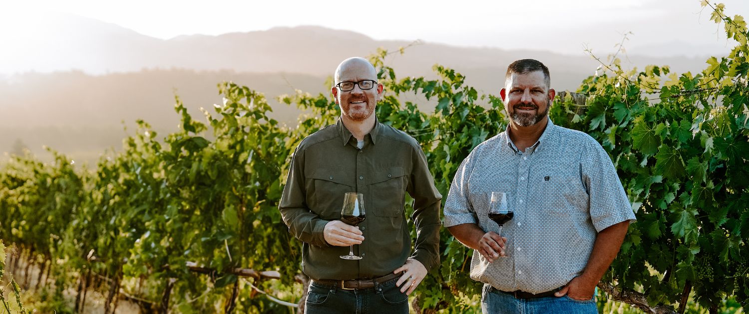 Andy and Ned standing in a vineyard with a wine glass.