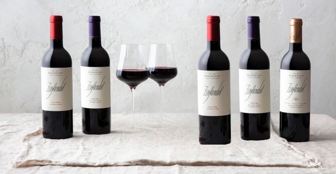 Five bottles of red wine are arranged in a row on a light cloth, with two glasses of red wine between the bottles in the center—capturing the elegance of a fine wine shop against a plain, light-colored wall.