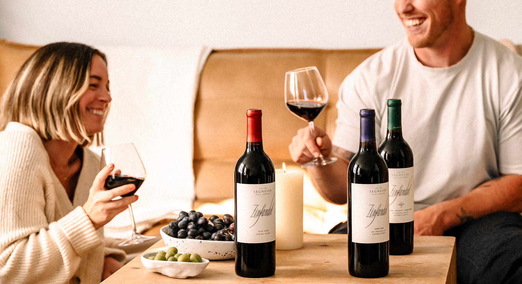 Two people sit on a couch, smiling and holding glasses of red wine. Three wine bottles, a candle, and bowls of grapes and olives are on the table in front of them, creating a cozy, relaxed atmosphere.