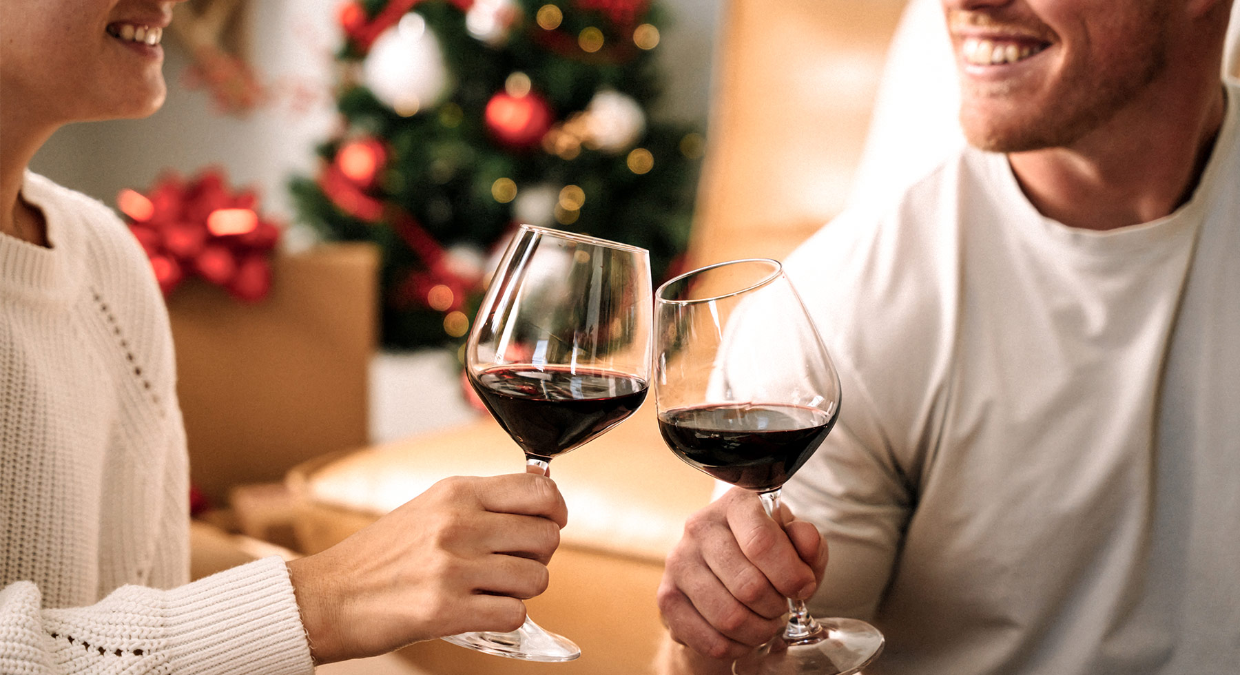 Two people smiling and clinking glasses of red wine together, with a decorated Christmas tree and wrapped presents in the background.