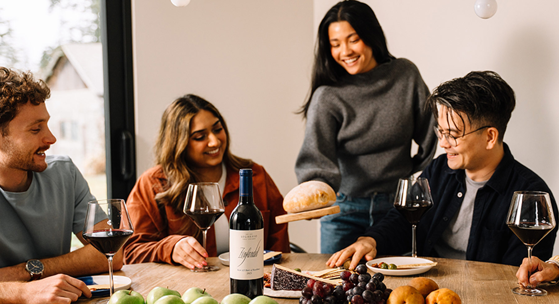 Four friends sit around a table with wine, fruit, and cheese, smiling and enjoying each others company. One person stands, serving bread, while the others sit and hold wine glasses. The atmosphere is warm and cheerful.