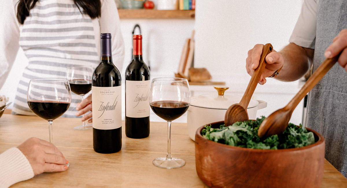 Two people enjoy wine in a kitchen; two glasses and two bottles of red wine are on the table, and someone is tossing a green salad in a wooden bowl with wooden utensils.
