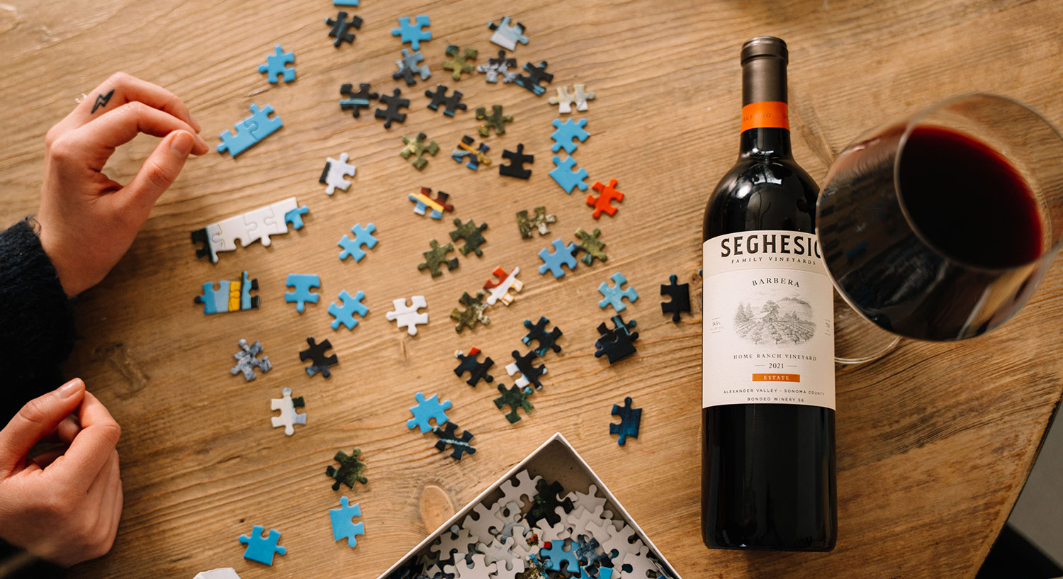 A person works on a scattered jigsaw puzzle next to a bottle of Seghesio wine and a glass of red wine on a wooden table.