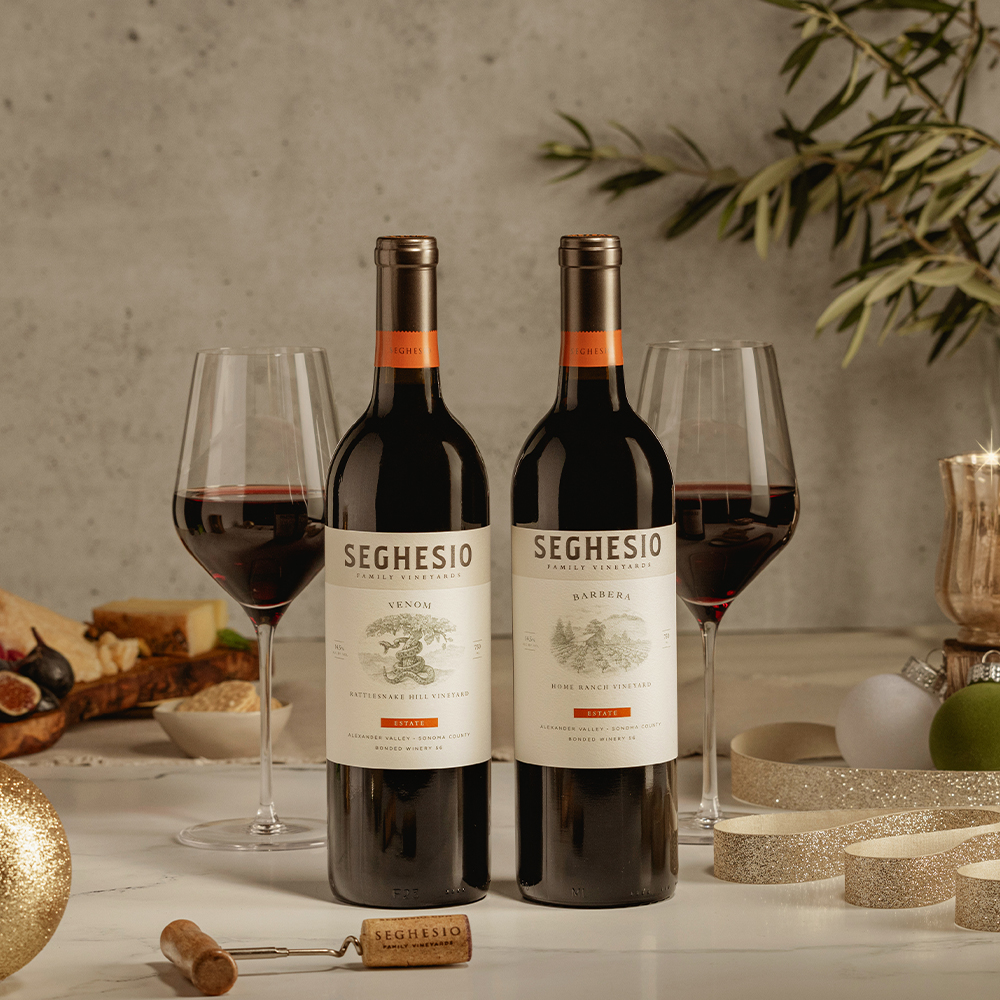 Two bottles from the Seghesio Favorites Wine Gift stand side by side on a table, with two filled wine glasses, a cork, ribbon, holiday ornaments, and a cheese platter in the background, creating a festive atmosphere.