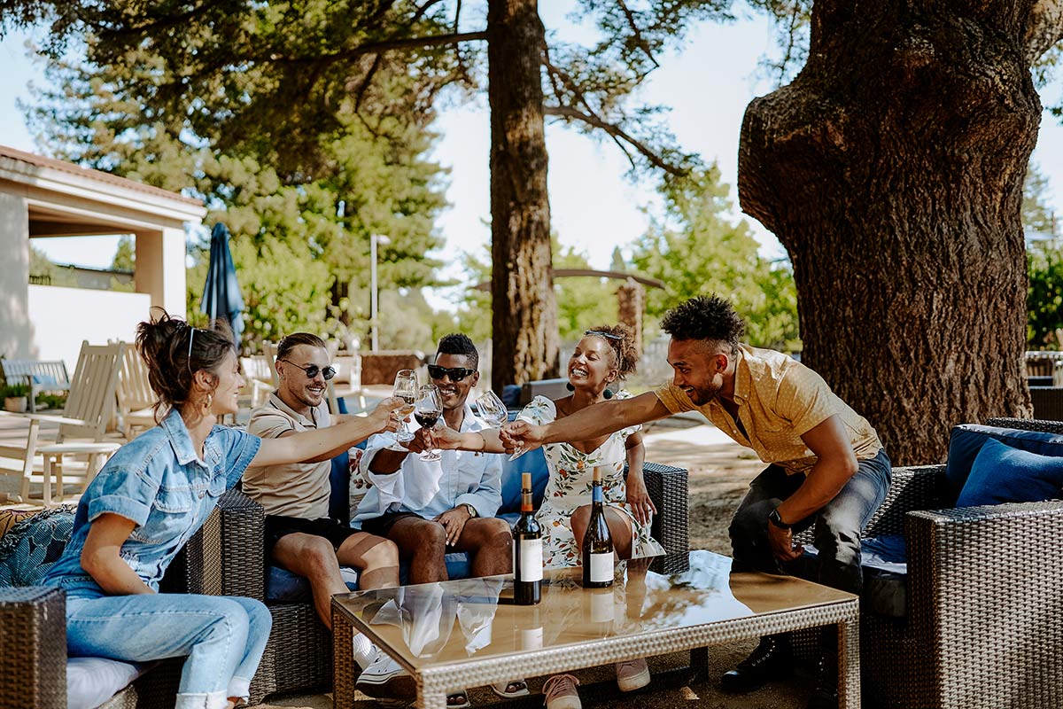 Five friends sit outside on wicker furniture, smiling and raising glasses in a toast around a glass-top table with wine bottles. Tall trees and sunny weather create a relaxed, cheerful atmosphere.