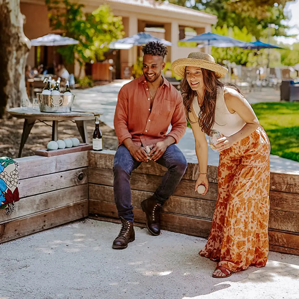 A smiling woman in a sunhat and floral skirt plays bocce ball while a man in a coral shirt and boots watches. They are outdoors on a sunny day, with drinks and bocce balls nearby on a wooden ledge.