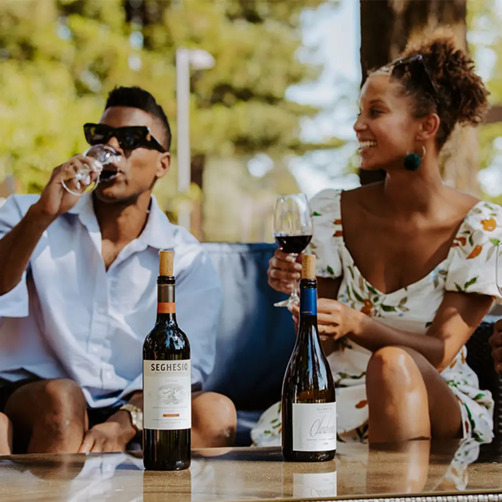 A man and woman sit outdoors on a sofa, enjoying glasses of wine. Two bottles of wine are on the table in front of them. They are smiling and surrounded by trees and sunlight.