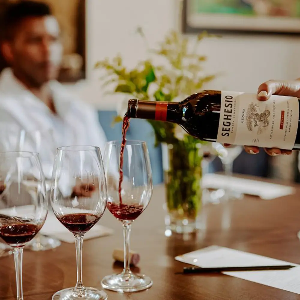 A hand pours red wine from a Seghesio wine bottle into glasses on a wooden table, with a blurred person in the background and a vase of flowers nearby.