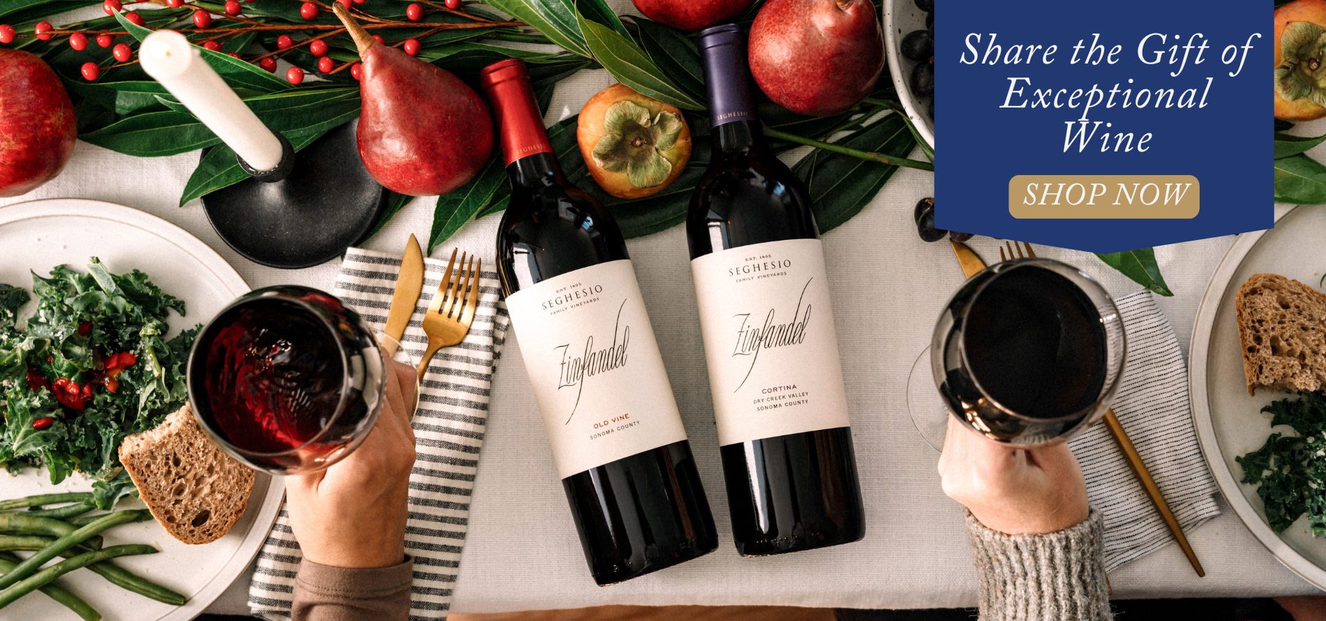 Two people toast with glasses of red wine over a festive table set with wine bottles, fruit, leafy greens, bread, and candles. A banner reads, “Share the Gift of Exceptional Wine from our tasting room—Shop Now.”.