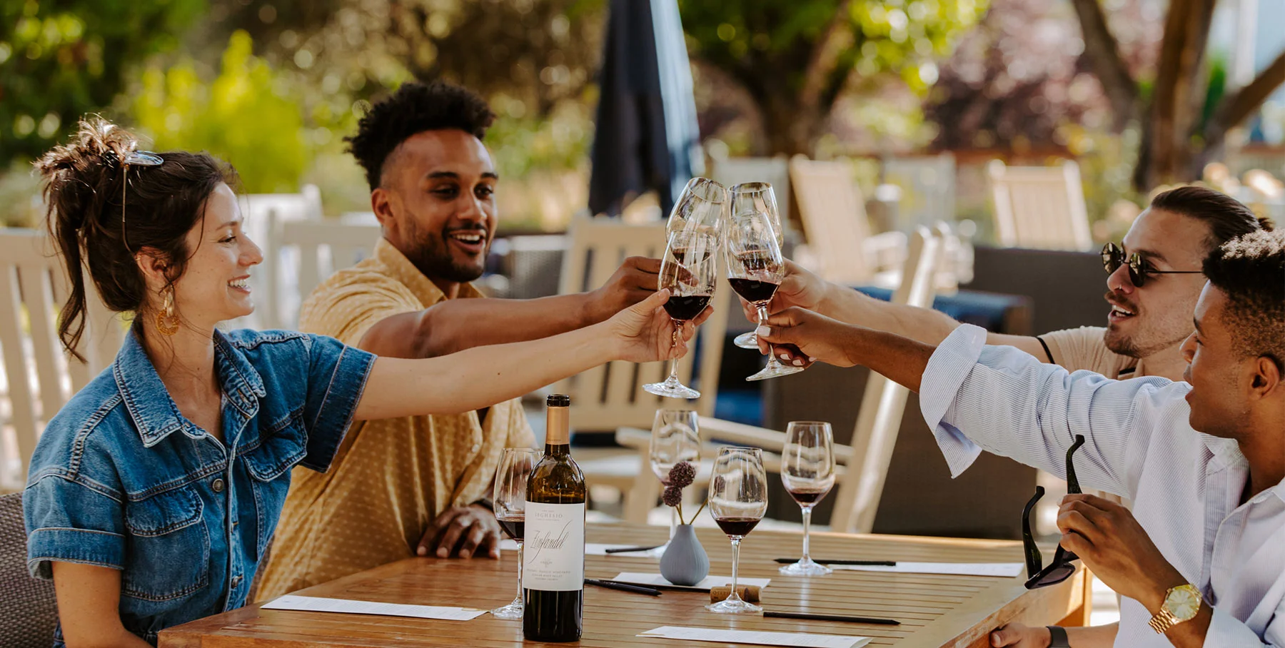 Four people sit at an outdoor tasting room table, clinking wine glasses in a cheerful toast. They smile and enjoy each other's company, surrounded by trees and sunlight, with a bottle of wine and glasses on the table.