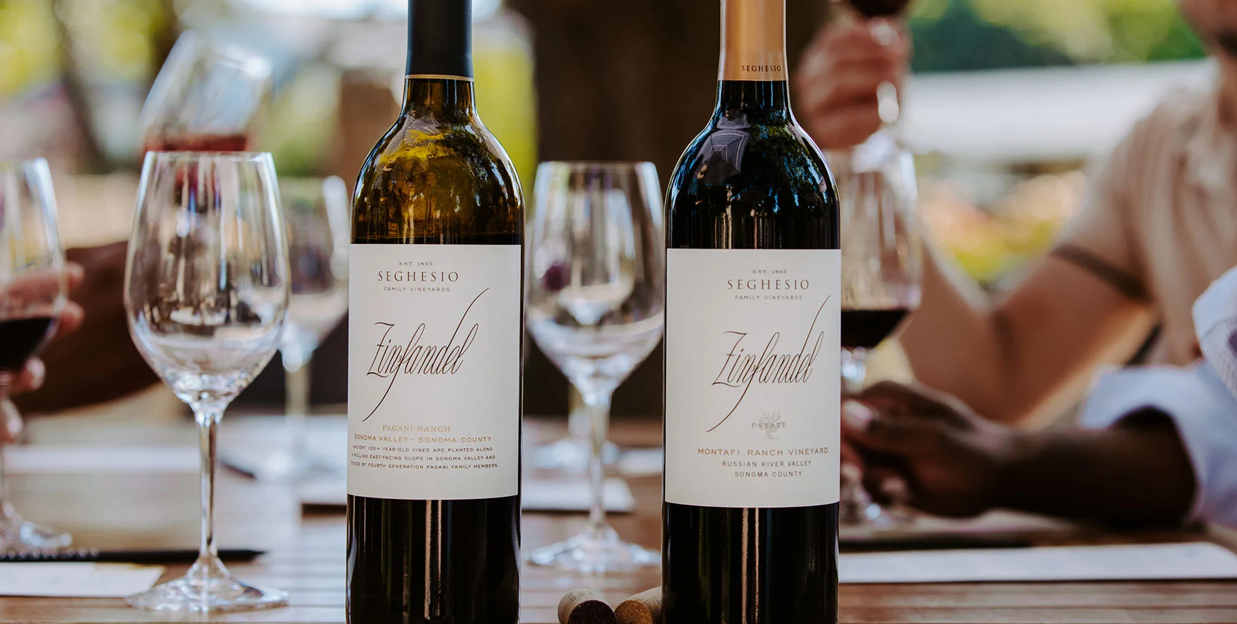 Two bottles of Seghesio Zinfandel wine sit on a wooden table outdoors, surrounded by wine glasses, corks, and people savoring a tasting room experience in the background.