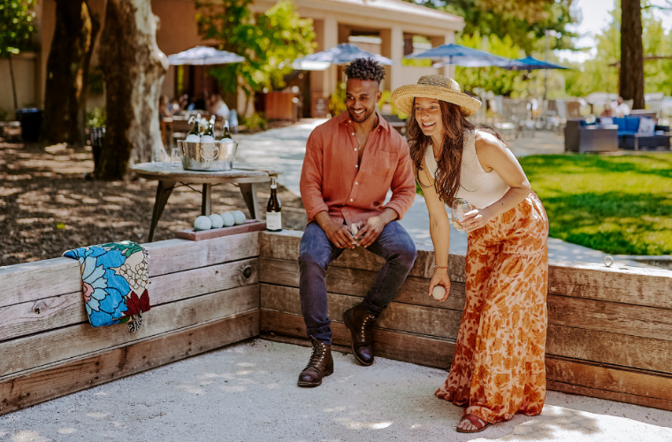 A woman in a straw hat and floral skirt plays bocce ball while smiling, as a man in a rust-colored shirt watches and sits nearby. They are outdoors near a table with drinks and a towel draped over the fence.