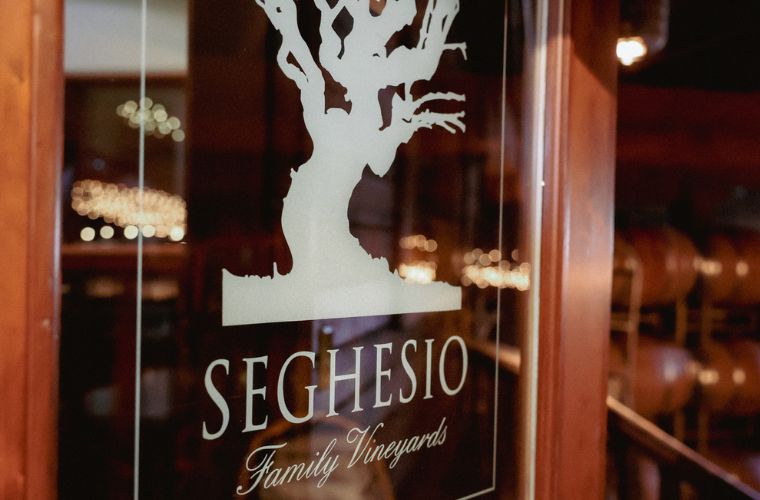 A glass door with a white tree silhouette and the words Seghesio Family Vineyards etched on it. Wine barrels are visible inside the warmly-lit room beyond the door.