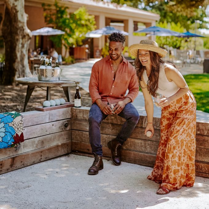 A man and woman play bocce ball outdoors on a sunny day. The woman, smiling, throws a ball as the man watches. Both are casually dressed, with trees, tables, umbrellas—and wine club newsletters—seen in the background.