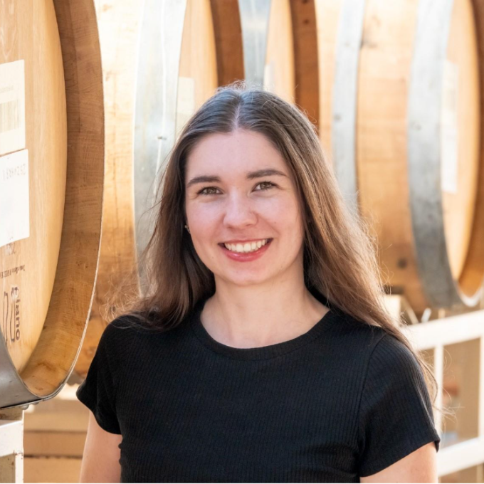 A woman with long brown hair and a black shirt smiles while standing in front of large wooden barrels, as if posing for wine club newsletters in a well-lit setting.