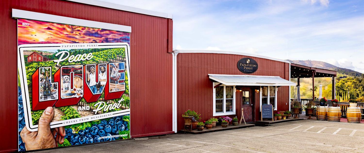 A red building with a large colorful mural reading “Peace, Love and Pies” shows berries and people’s hands. The entrance has a “Parkerville Berry” sign, several barrels outside, and mountains in the background—perfect for wine club newsletters.