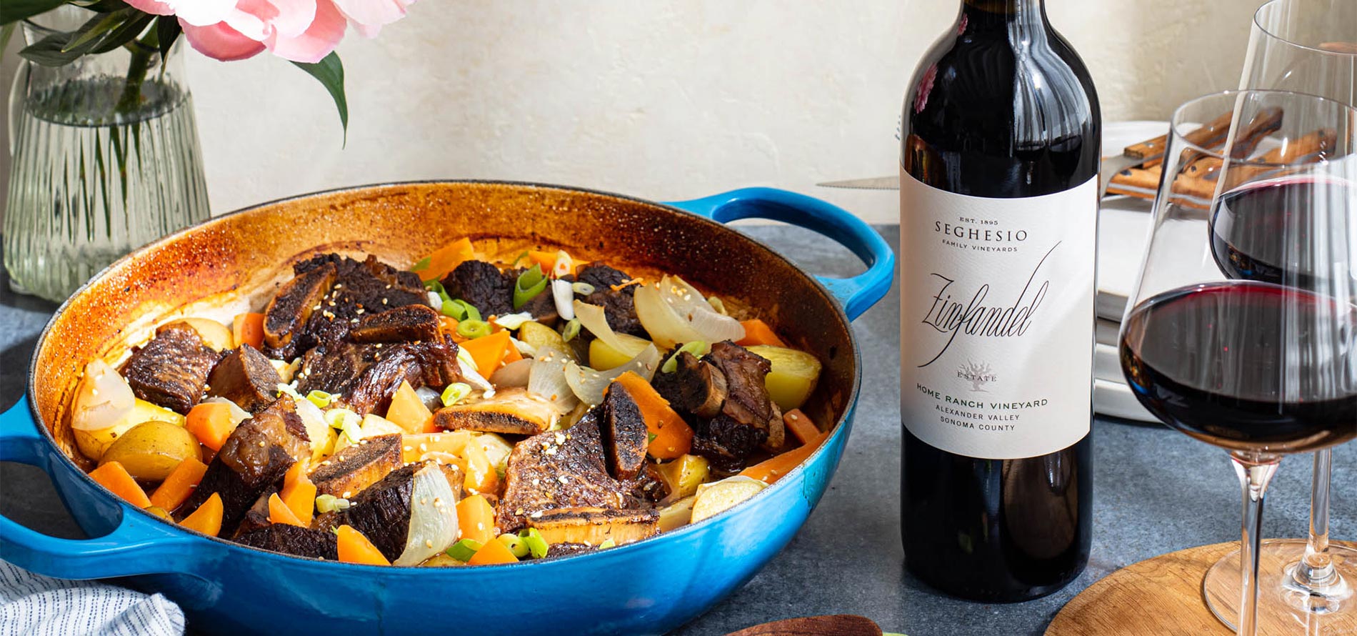 A blue Dutch oven filled with braised beef, carrots, onions, and potatoes is set on a table next to two glasses and a bottle of Seghesio Zinfandel red wine. A fork and napkin are in the background.