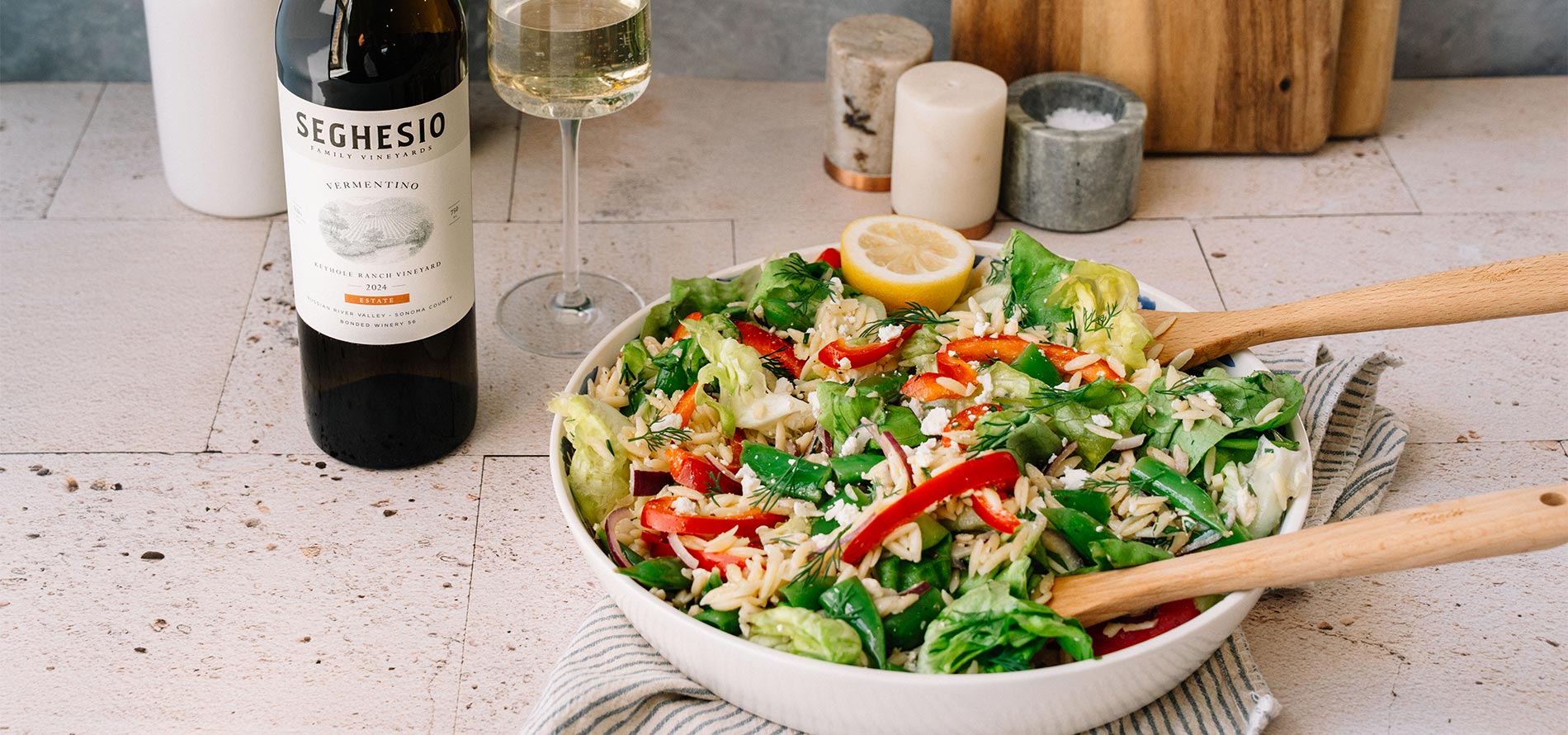A fresh salad with orzo, lettuce, bell peppers, and herbs sits in a white bowl with wooden utensils. A bottle of Seghesio wine, a glass of white wine, and candles are on the counter nearby.
