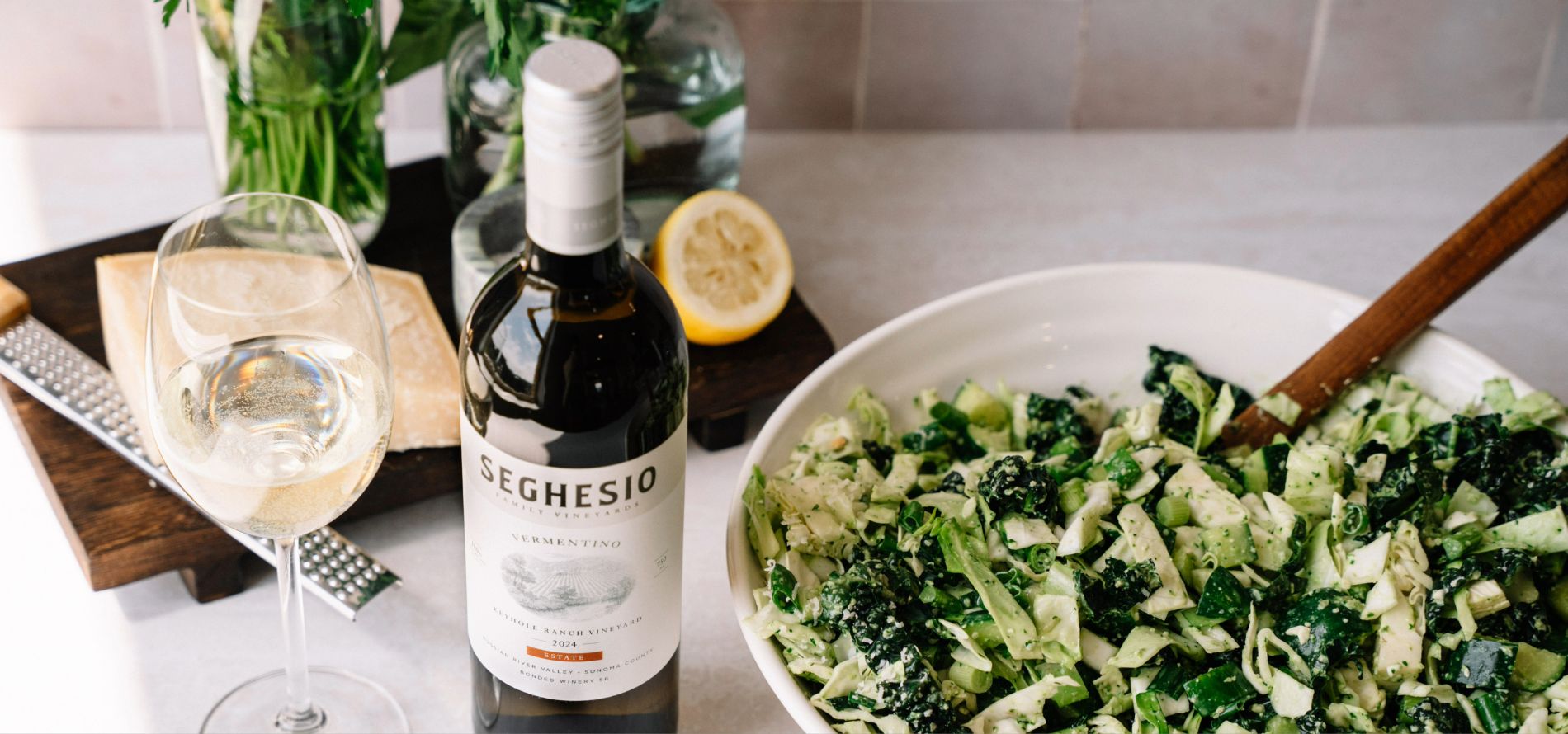 A bottle of Seghesio Vermentino wine, a filled wine glass, a bowl of green salad, Crispy Sage with Brown Butter Mushroom Ravioli, a halved lemon, and a vase of flowers are arranged on a white countertop.