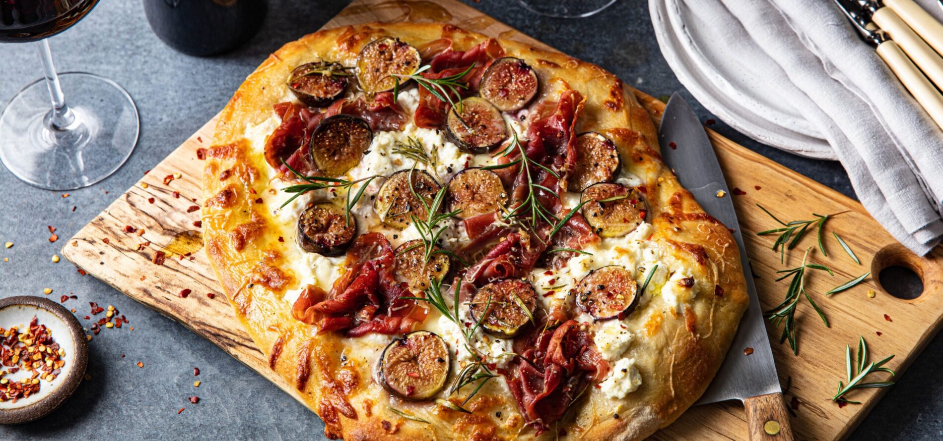 A rectangular flatbread topped with sliced figs, prosciutto, creamy cheese, and rosemary, served on a wooden board with a knife. Wine glasses, a stack of plates, cutlery, and a small bowl of chili flakes are nearby.