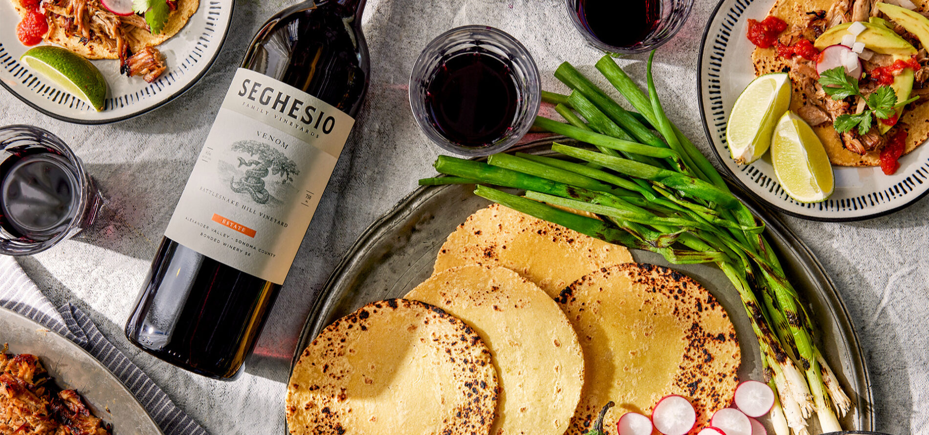 A bottle of Seghesio red wine lies on a table with corn tortillas, grilled green onions, sliced radishes, lime wedges, and plates of tacos. Glasses of red wine accompany the meal.