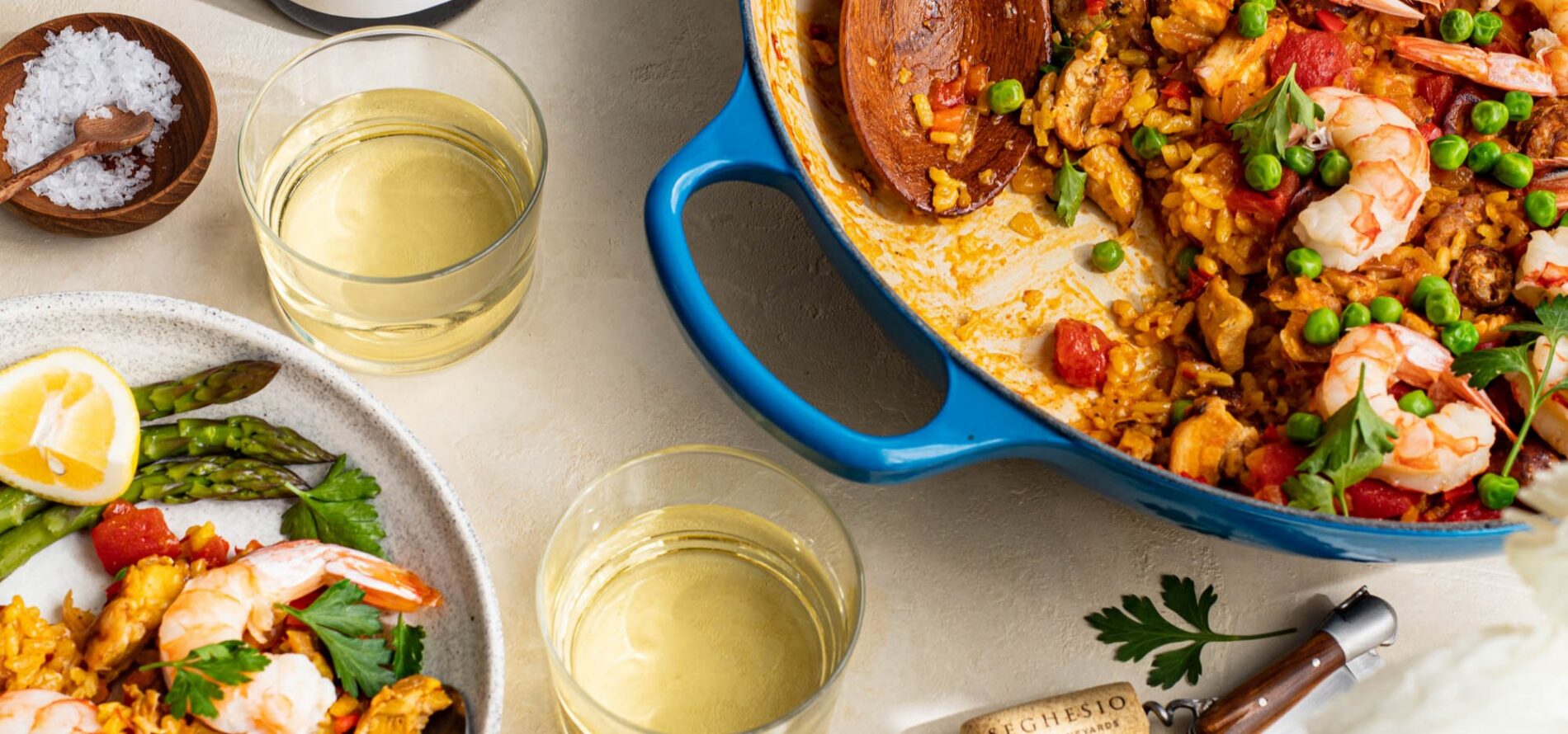A blue pan filled with shrimp and vegetable paella sits beside two glasses of white wine, a plate with paella and asparagus, a bowl of salt, and a wine cork on a light table.