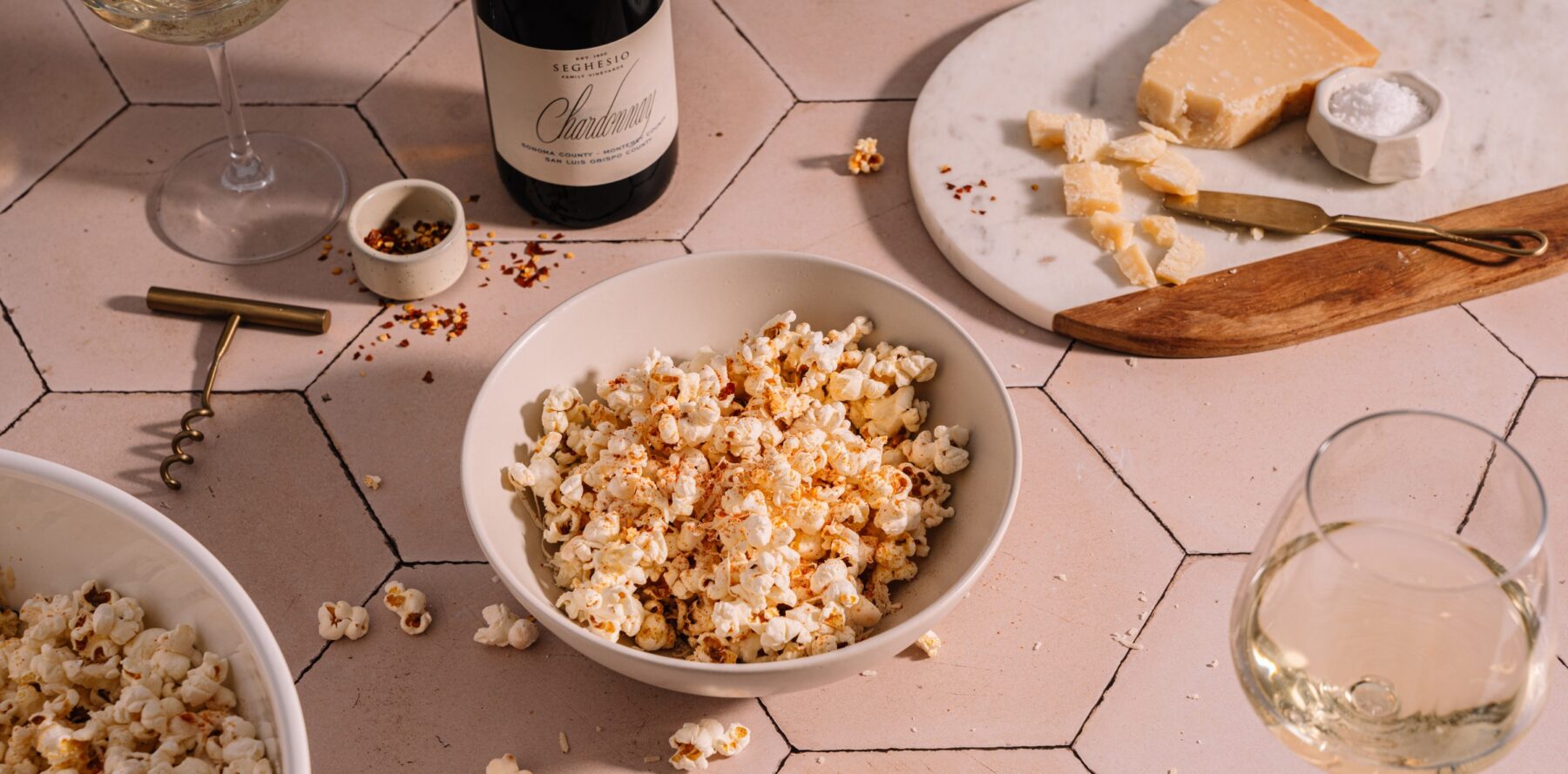 A bowl of popcorn sprinkled with seasoning sits on a tiled surface, surrounded by glasses of white wine, a bottle of wine, a corkscrew, and a marble board with chunks of cheese and a small dish of salt.