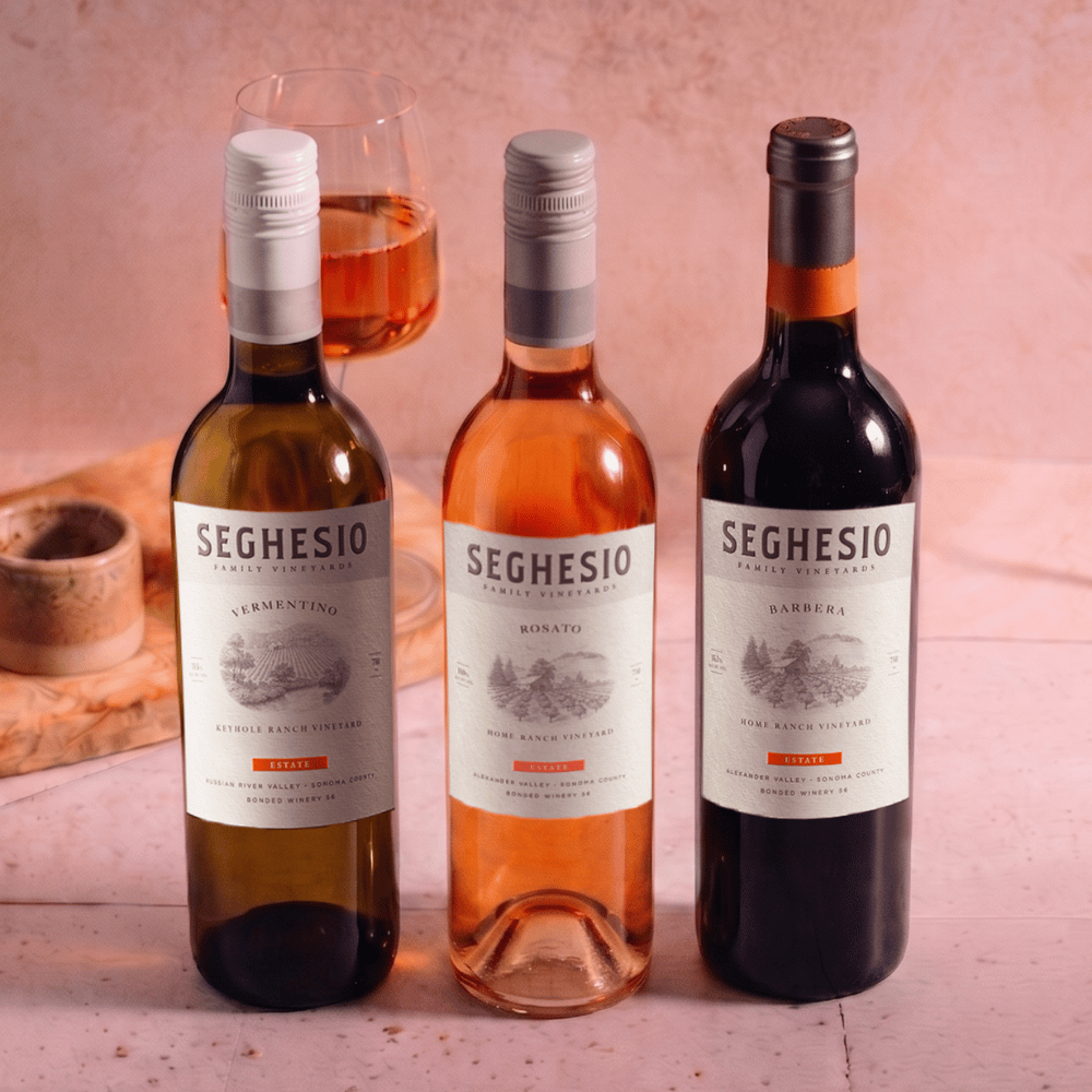 Three bottles of Seghesio wine—white, rosé, and red—are displayed on a pink surface with a filled wine glass and a ceramic dish in the background. The labels are clearly visible, showing the wine varietals.