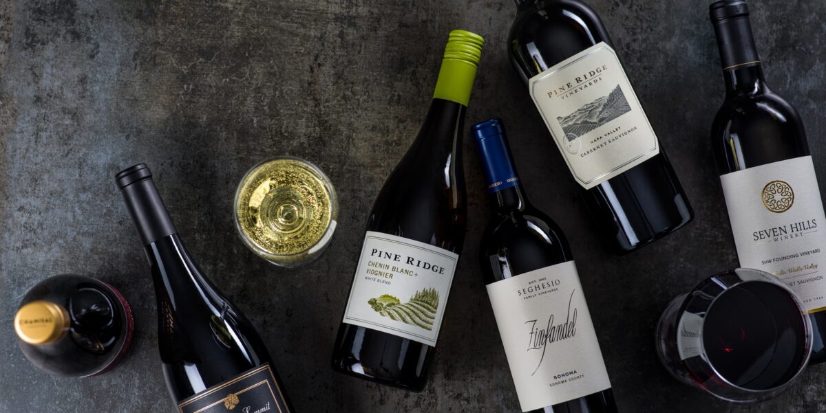 A variety of red and white wine bottles are arranged on a dark surface, with two filled wine glasses—one with white wine, one with red—placed among the bottles.