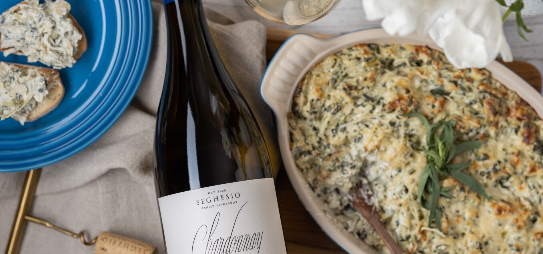 A bottle of Seghesio Chardonnay lies next to a dish of baked cheesy spinach dip, a blue plate with bread and dip, a wine glass, and a corkscrew on a wooden surface.
