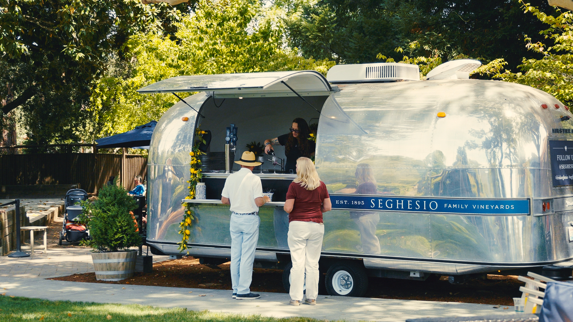 A man and woman stand at the counter of a shiny silver food trailer labeled Seghesio Family Vineyards in a park setting, being served by a person inside. Tall trees and outdoor seating are visible in the background.