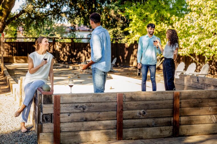 Four people gather outdoors by a wooden bocce court, chatting and holding drinks. One woman sits on the edge, while three others stand nearby under sunny trees, enjoying a relaxed, social atmosphere.
