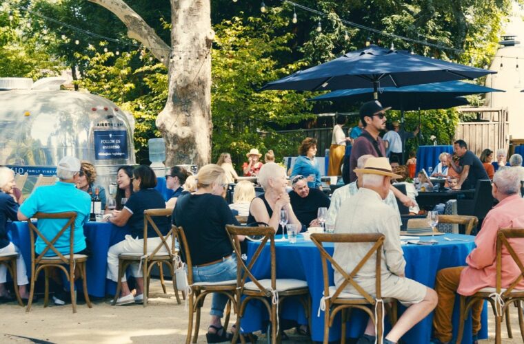 People are seated at round tables with blue tablecloths outdoors, eating and talking. Some stand or walk nearby. The area is shaded by trees and umbrellas, with a food truck or trailer in the background.
