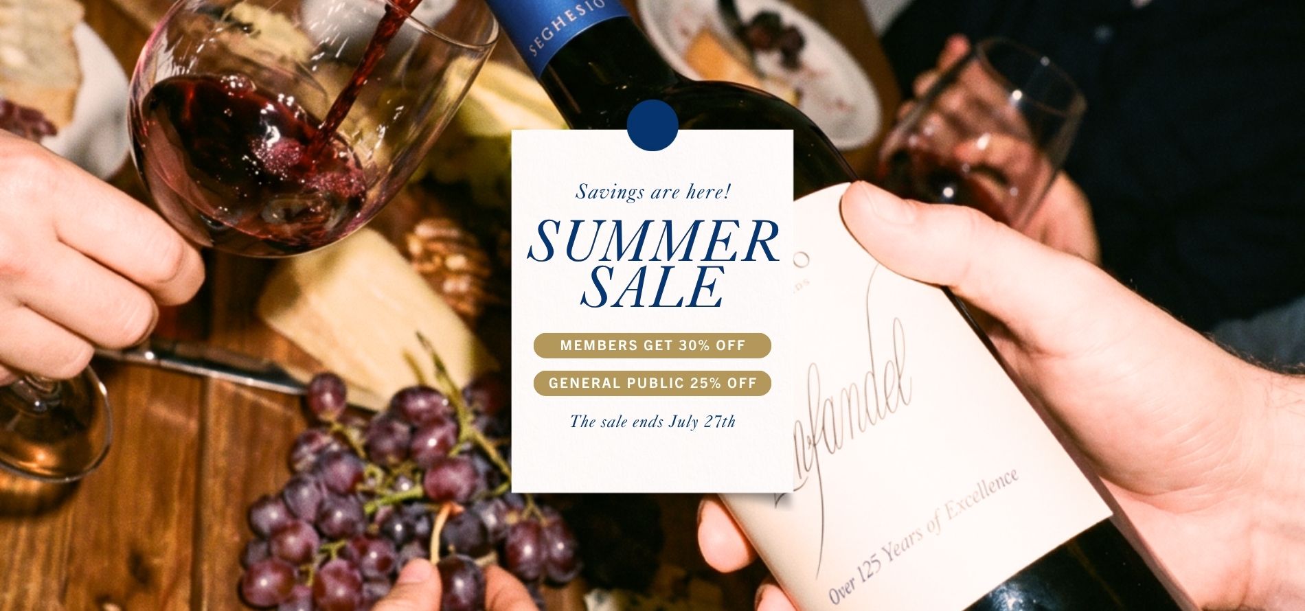 Close-up of people pouring and drinking red wine in a tasting room at a table with grapes and cheese. A sign reads “Summer Sale: Members get 30% off, General Public 25% off. The sale ends July 27th.”.