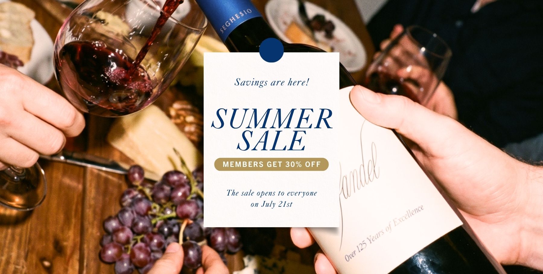 A hand holds a wine bottle displaying a “Summer Sale” sign in the tasting room; others raise wine glasses over a table with grapes and food. The sign offers 30% off for members and notes the sale opens July 21st.