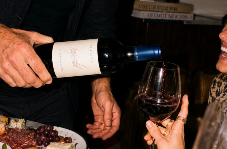A person pours red wine from a bottle into a glass held by another person, with a charcuterie board and books visible in the background.