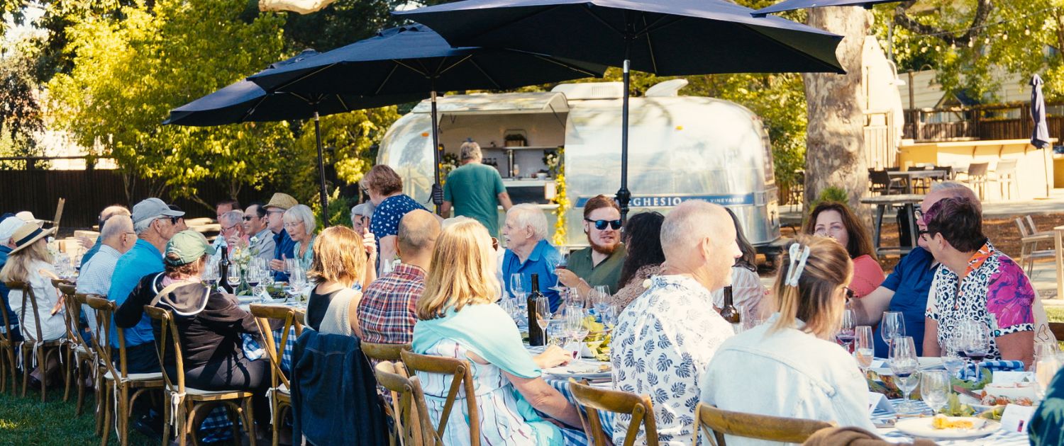 A group of people sit at a long outdoor table enjoying a meal together under large black umbrellas, with a silver food trailer in the background and trees providing shade—like a lively gathering straight out of wine club newsletters.