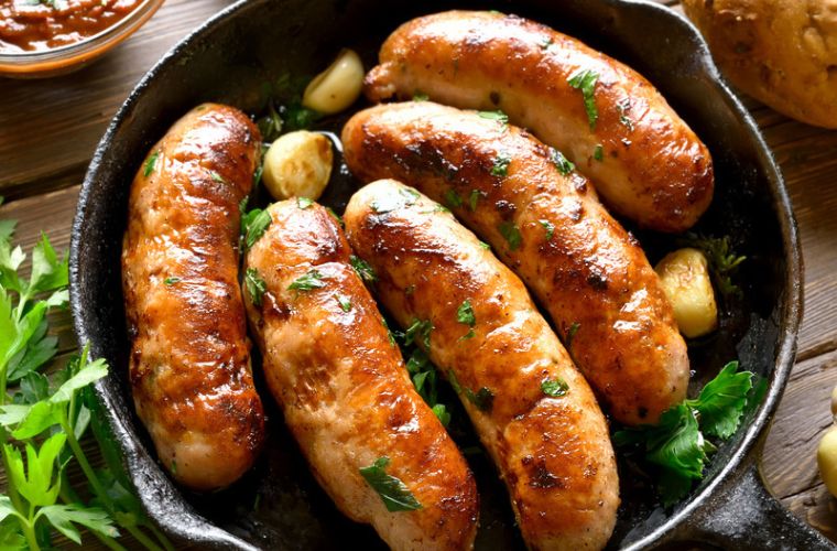 Four cooked sausages with herbs and garlic cloves in a cast-iron skillet.